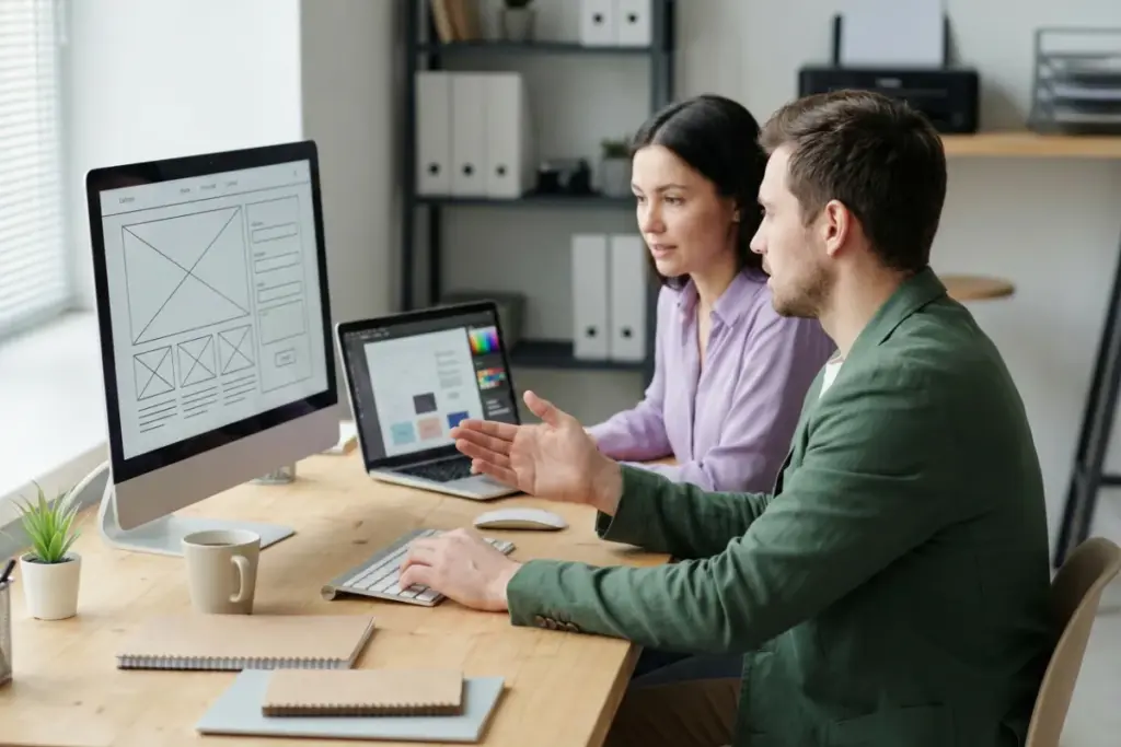 A man and a woman working together at a desk with monitors and a laptop, illustrating realistic front-end and back-end website development in a professional office setting.